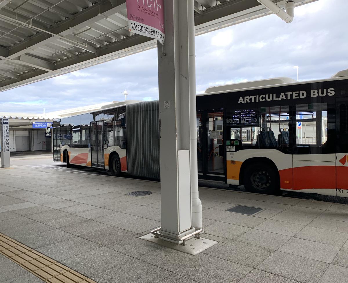 第 2 航廈門口有頂棚的人行道旁，停著一輛白橘配色、車身寫「ARTICULATED BUS」的南海連節巴士；左邊立柱寫「無料連絡バスおりば Free Shuttle Buses Drop-off Area」，柱子上掛著寫「欢迎来到日本」的廣告旗