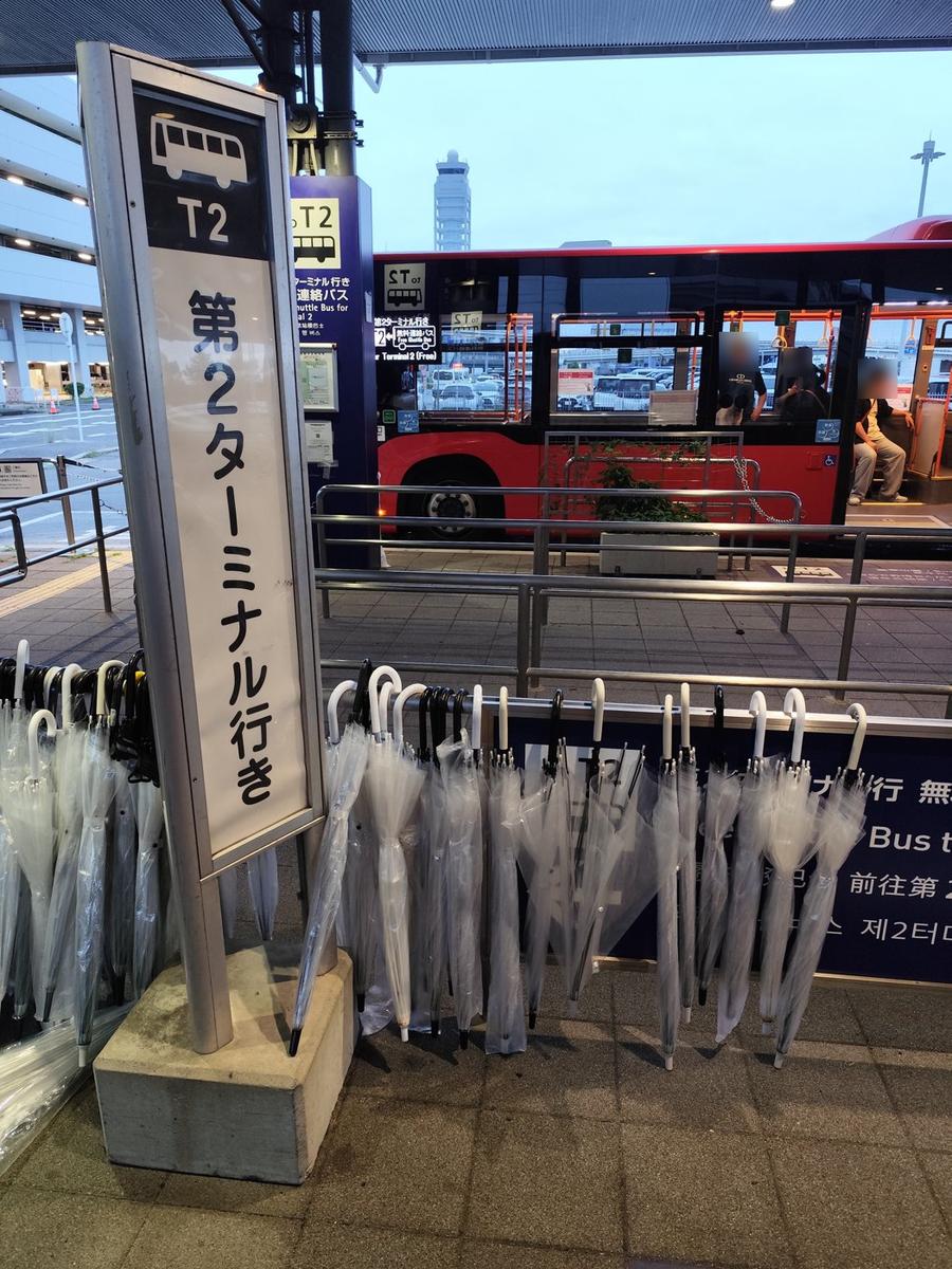 Aeroplaza 1 樓有頂棚的巴士站，前方直立站牌寫「T2 第2ターミナル行き」，站牌下掛著一排透明雨傘；後面停著紅色的連絡巴士，車窗上貼著「T2 第2ターミナル行 無料連絡バス Free Shuttle Bus」，遠方是關西機場的管制塔