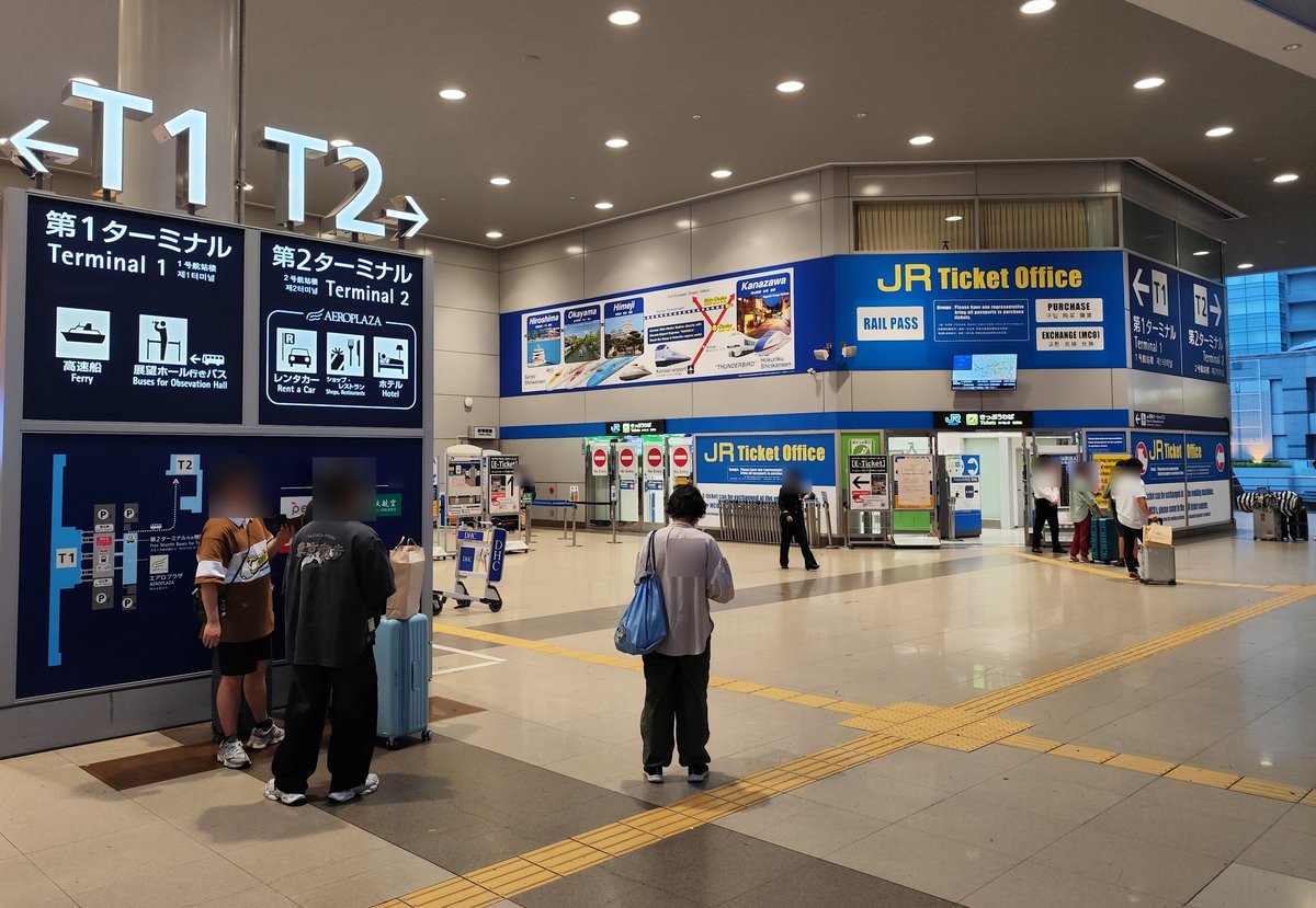 關西空港站剪票口外的大廳，左邊大型立牌頂端寫「←T1」「T2→」，左半是第 1 航廈（高速船、展望ホール行きバス），右半是第 2 航廈，下面框著 AEROPLAZA 與租車、商店餐廳、飯店圖示；右後方是藍色的 JR Ticket Office 與剪票口