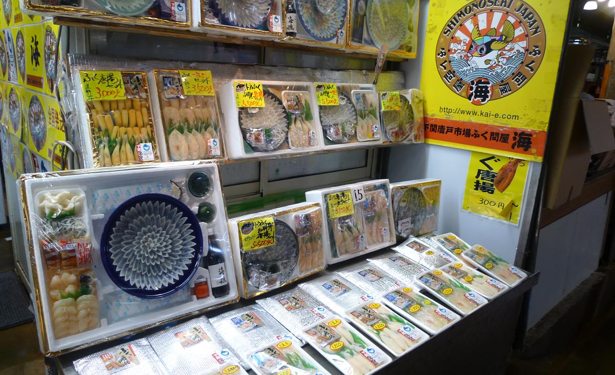唐戸市場河豚批發店「海」的店頭:盒裝的河豚刺身(薄片排成菊花狀的藍色大盤)、河豚皮、河豚唐揚,貼著黃色價格牌,右邊是黃色店招