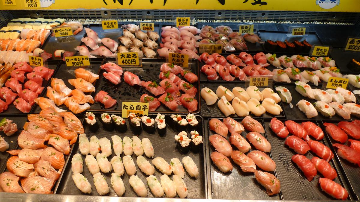 壽司街攤位的黑色托盤上排滿單貫握壽司:鮭魚、鮪魚大腹、白肉魚、鱈魚白子軍艦,每排插著黃色價格牌