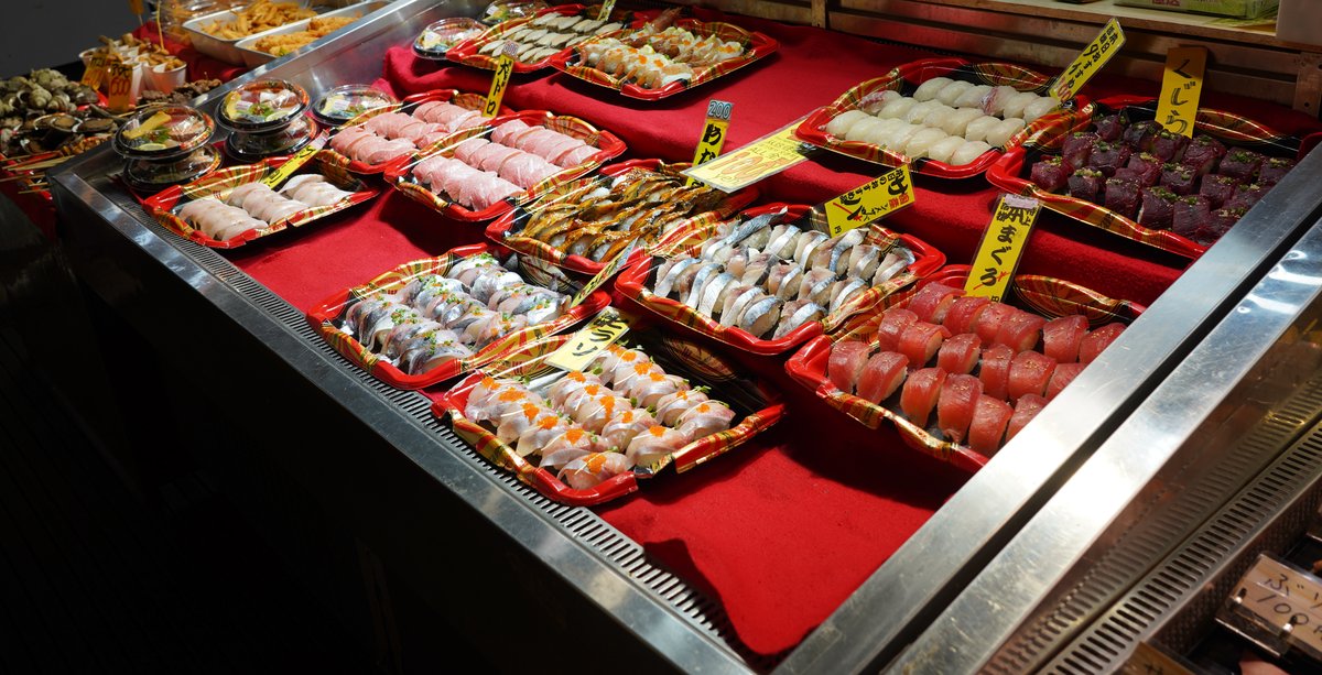 唐戸市場 1 樓攤位的紅色展示台上排滿一盤盤握壽司,鮪魚、白肉魚、青背魚各一盤,插著黃色手寫品名牌