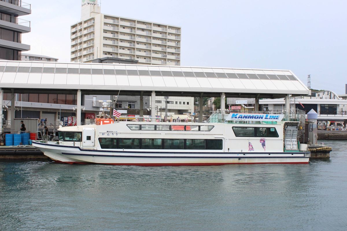 下關側的唐戸棧橋:白色大屋頂的浮動碼頭旁停著一艘船身寫「がんりう」、掛 KANMON LINE 招牌的白色客船,背後是公寓大樓