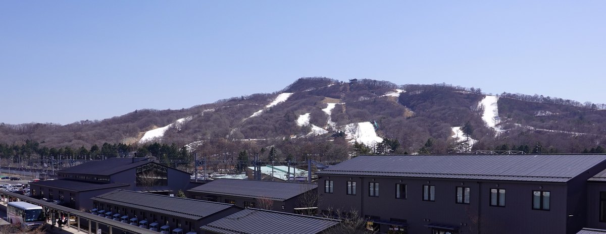 從輕井澤站的天橋看輕井澤王子滑雪場整座山：褐色的落葉樹林裡有七、八道白色雪道，前景是站前深色屋頂的商場與鐵路架線
