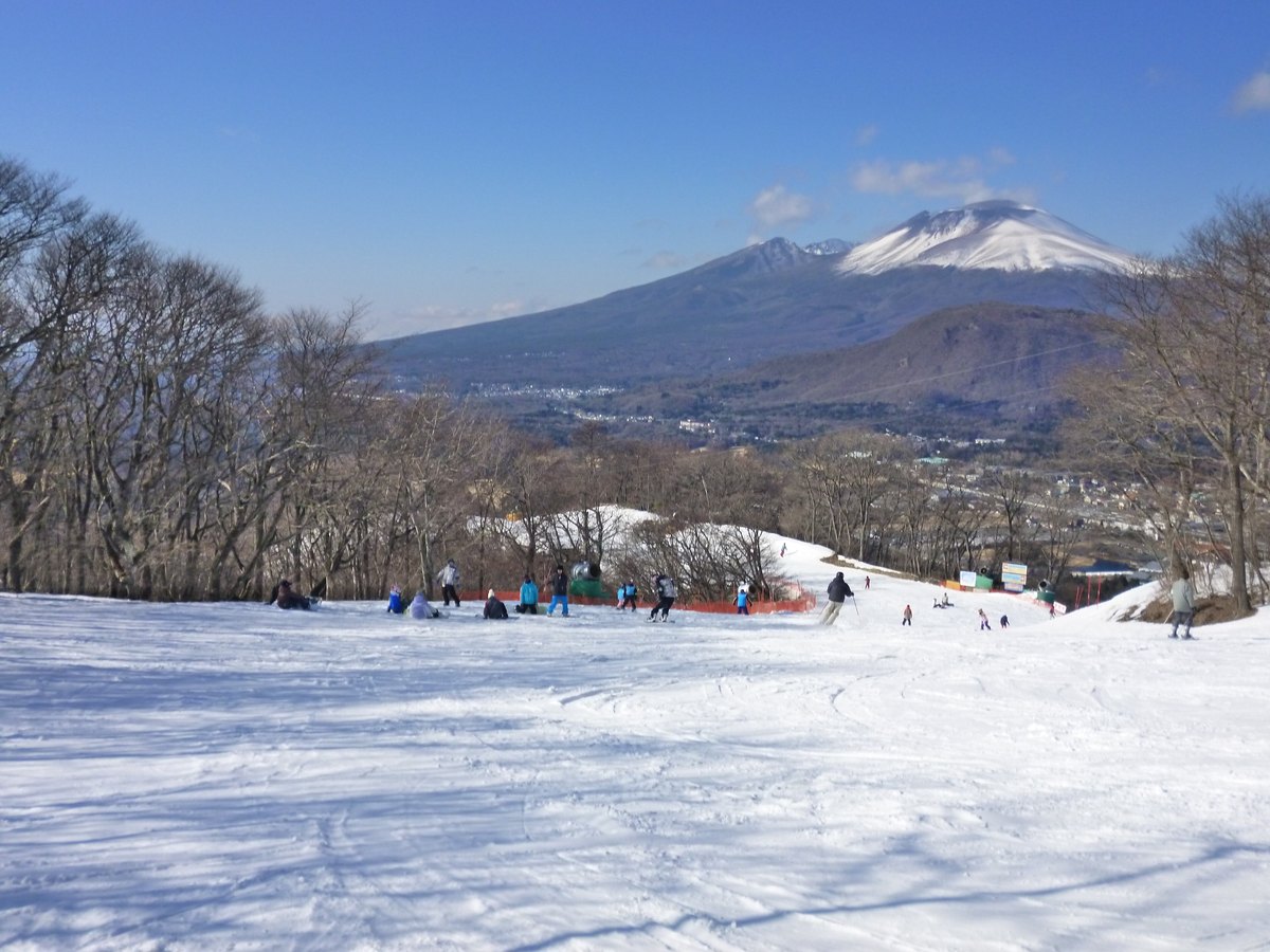 從輕井澤王子滑雪場的雪道往下看：前景是壓過的雪面，幾位滑雪客坐在坡邊，橘色護網旁有一台綠色造雪機，遠方是山頂積雪的淺間山與山腳的輕井澤市街
