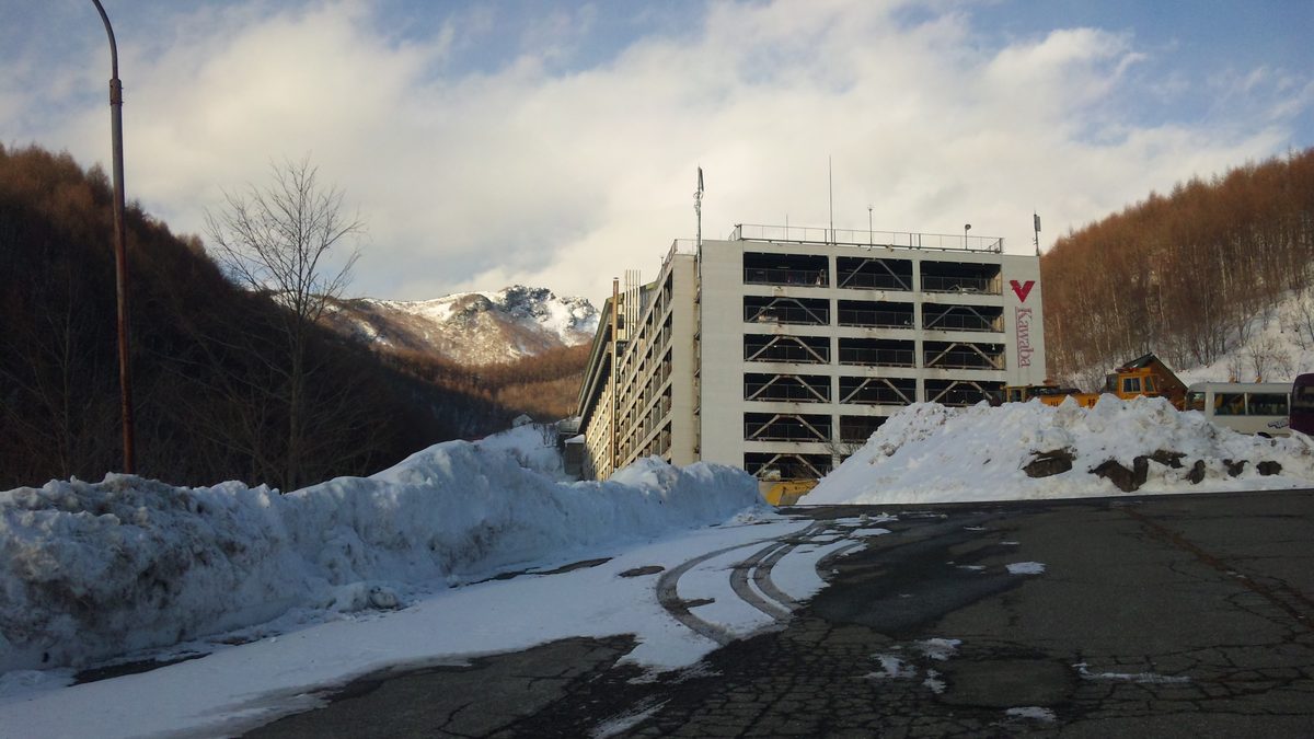 川場滑雪場的立體停車場:多層的白色停車樓,外牆有紅色 V 字標誌和直寫的 Kawaba 字樣,前方道路兩側堆著剷起來的雪,右側停著一輛小巴與黃色工程車,背後是落葉松山坡與覆雪的岩峰