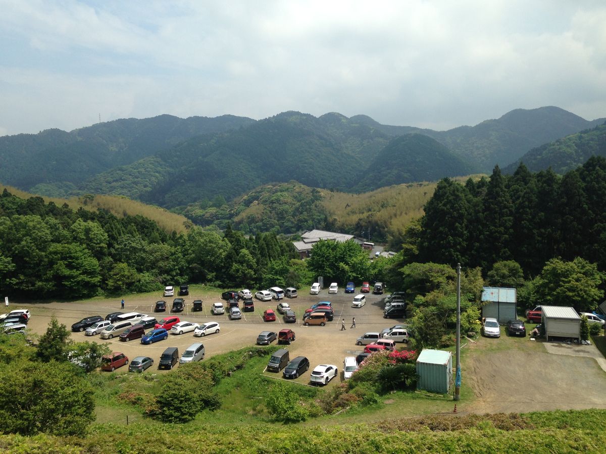 從高處俯瞰河內藤園的停車場：一片沒有鋪柏油的土黃色空地上停著數十台車，白線直接畫在土面上，場地被樹林包住，後方是一棟灰瓦屋頂的建築與層層疊疊的綠色山稜