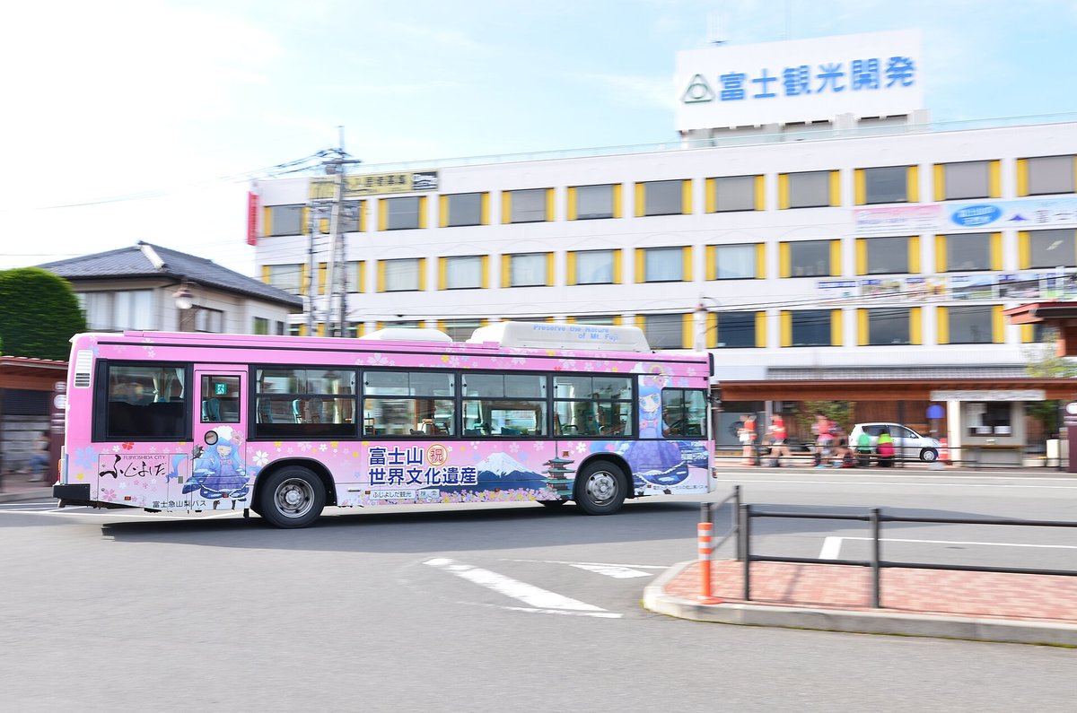 河口湖站前廣場對面一棟白色四層樓,窗框漆成黃色,屋頂立著藍字「富士観光開発」的大招牌;樓前一台印著富士山與「富士山 世界文化遺産」字樣的粉紅色巴士開過,畫面因追焦而背景帶點動態模糊;2015 年 7 月攝