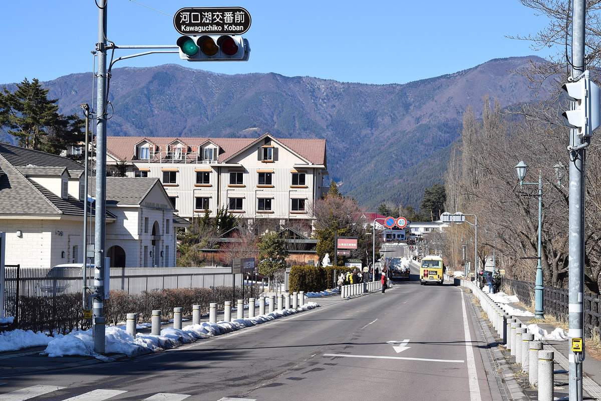 號誌牌寫著「河口湖交番前」的路口:車道是乾的,兩側人行道邊堆著殘雪,左側是山梨寶石博物館的白色建築,右側一排落葉的白楊,遠處一輛黃色巴士