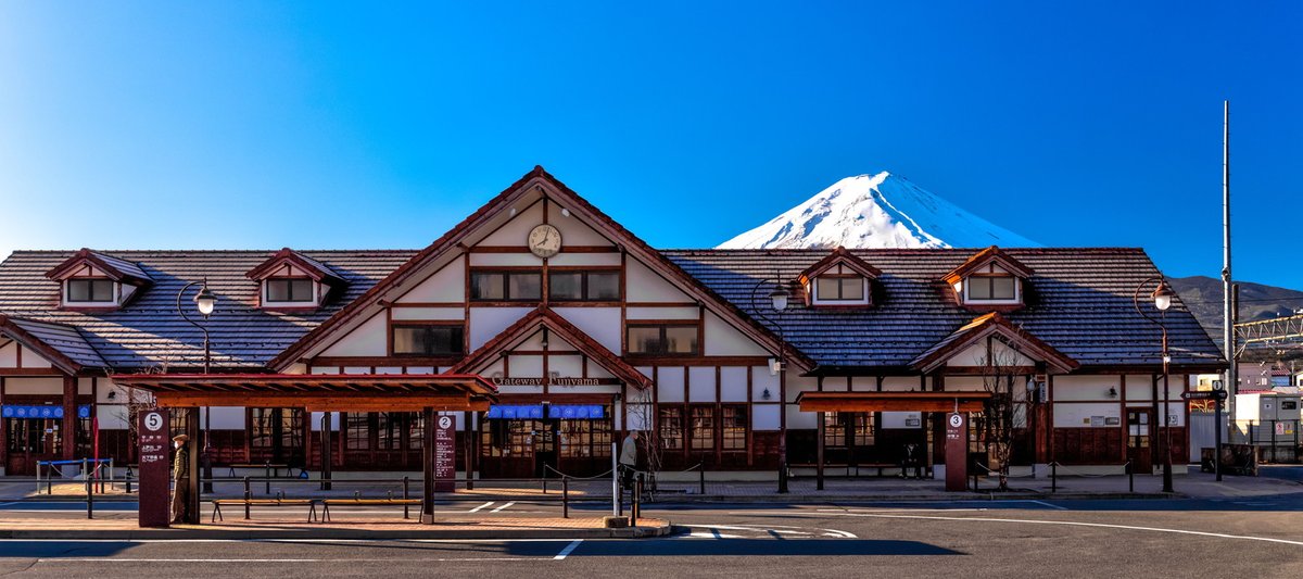 河口湖站站舍正面,屋頂後方露出山頂積雪的富士山;站前廣場有幾座編號的深紅色巴士站柱與候車棚,看得到 2、3、5 號