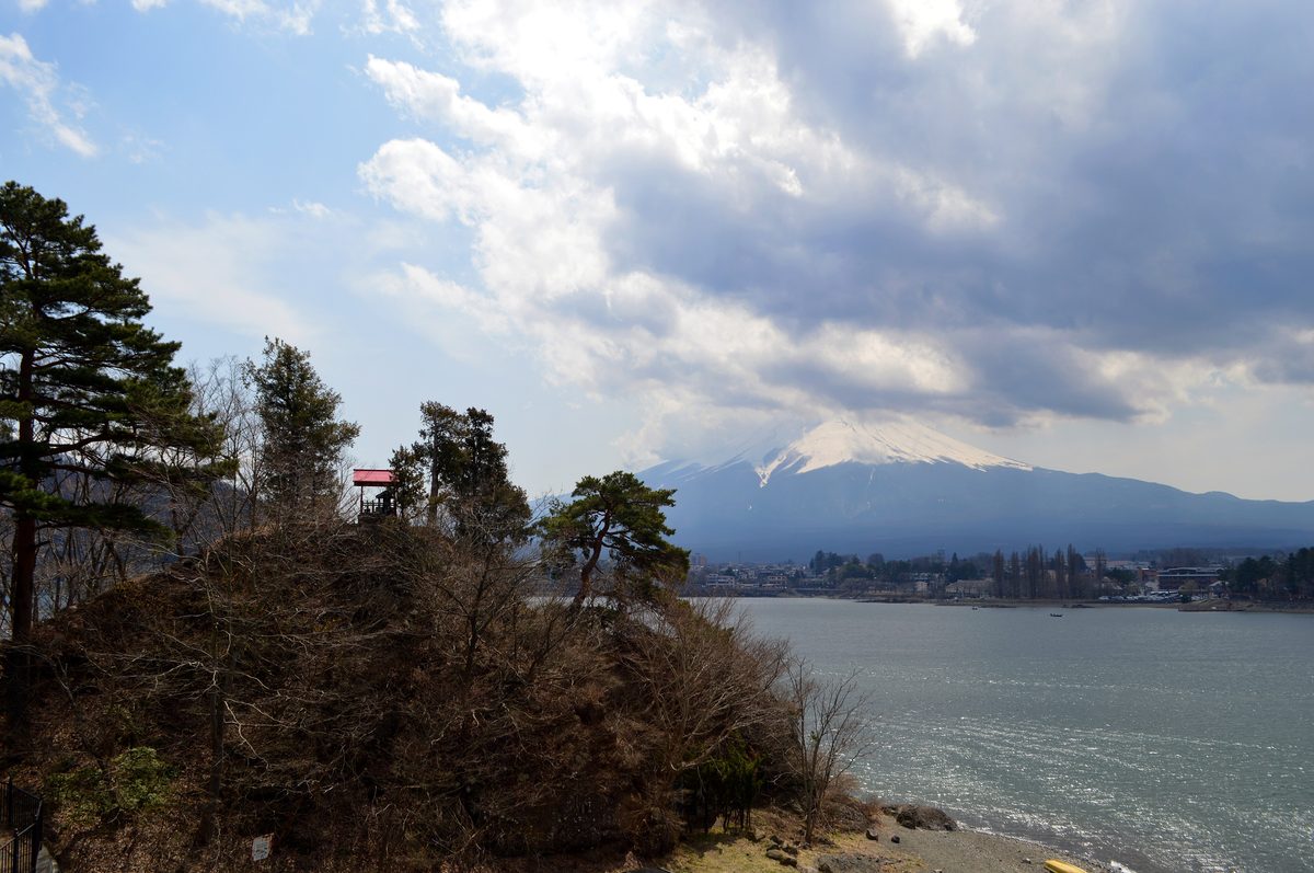 從湖邊高處看產屋ヶ崎:長著松樹的小岬角伸進河口湖,林間有一座紅色屋頂的小建築,右後方隔著湖是山頂積雪、上半被雲蓋住的富士山