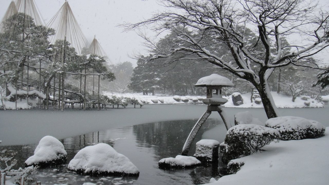 下雪中的兼六園霞ヶ池：地面、石頭與樹枝都覆著一層白雪，畫面中央是徽軫燈籠，左後方可以看到幾座雪吊的尖頂，空中還有正在飄落的雪花