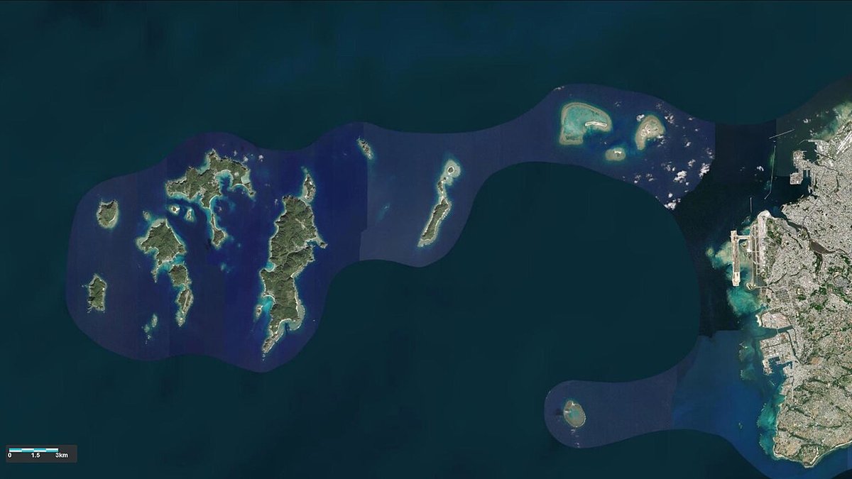Satellite view of the Kerama Islands, a scatter of dark green islands on deep blue water with a paler blue boundary shading drawn around them, southern Okinawa Island and Naha at the right edge, a 0 to 3 km scale bar bottom left