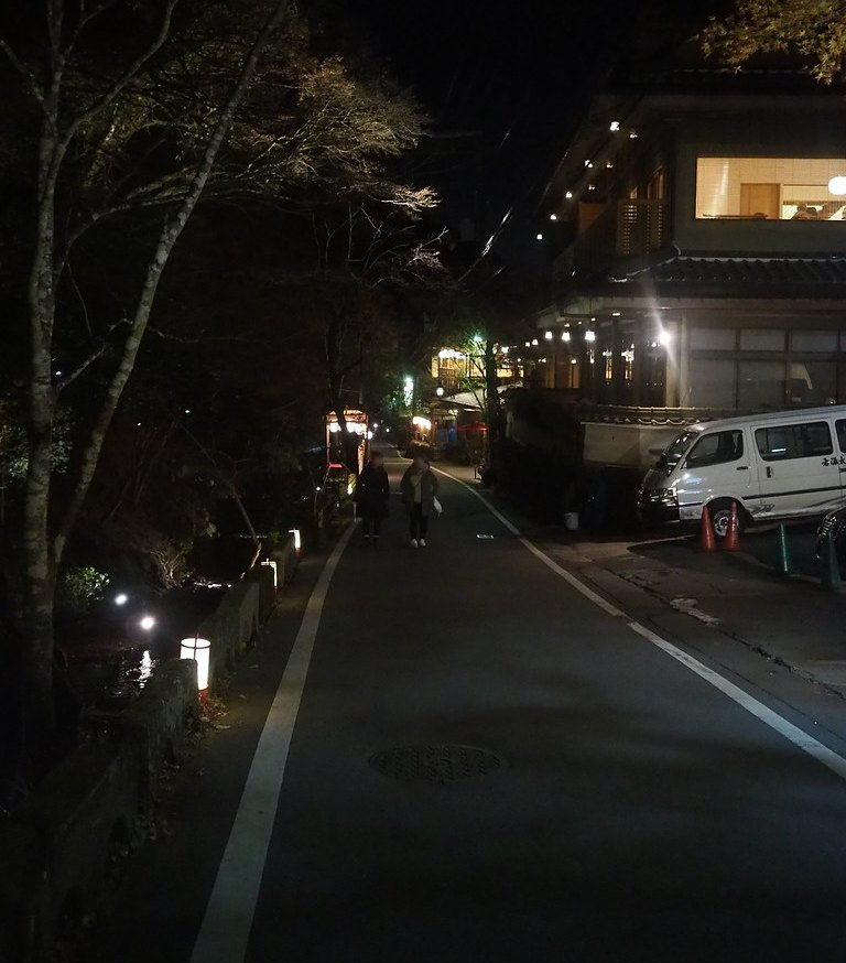夜間的貴船車道：柏油路兩側只畫白線、沒有另外隔開的人行道，兩個行人走在車道上，右邊是亮著燈的旅館與一台停著的白色廂型車，左邊矮牆上放著幾盞行燈，再過去是暗的溪谷