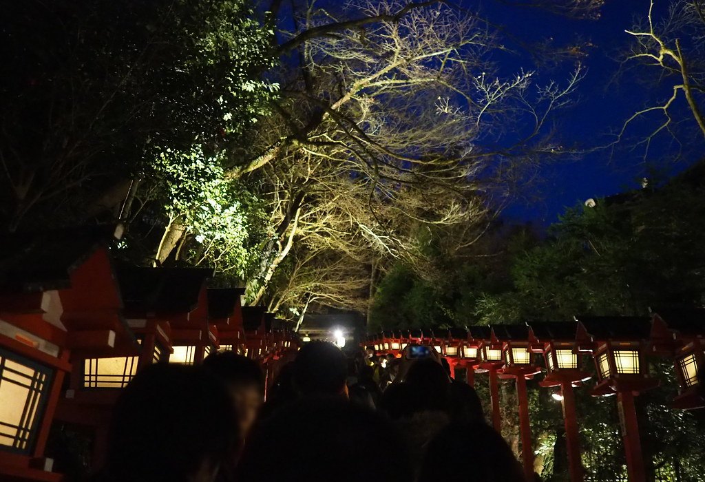 夜間的貴船神社本宮表參道：石階兩側的紅色春日燈籠亮著暖黃的燈，上方的樹被打成黃白色、枝上幾乎沒有葉子，天空是深藍色，石階上排滿人的背影