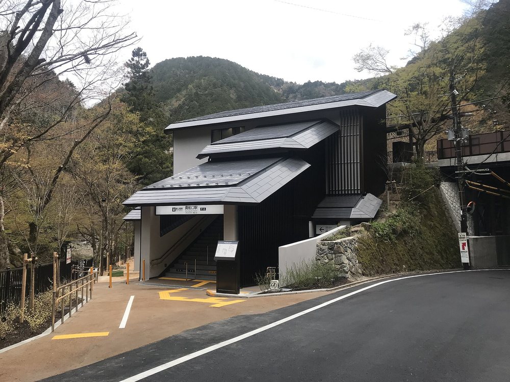 叡山電車貴船口站的站房：深灰屋頂、黑色格柵外牆的兩層建築，入口上方白底站名牌寫「貴船口駅 E16」，一道樓梯通往上層月台，站前是車道，周圍是山，樹還沒長葉