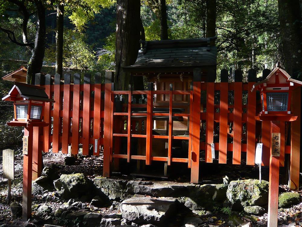 貴船神社結社：石頭基座上的朱紅色木柵圍著一座小社殿，兩側各一盞紅色春日燈籠，後方是高大的杉樹與轉黃的樹葉，陽光斜照