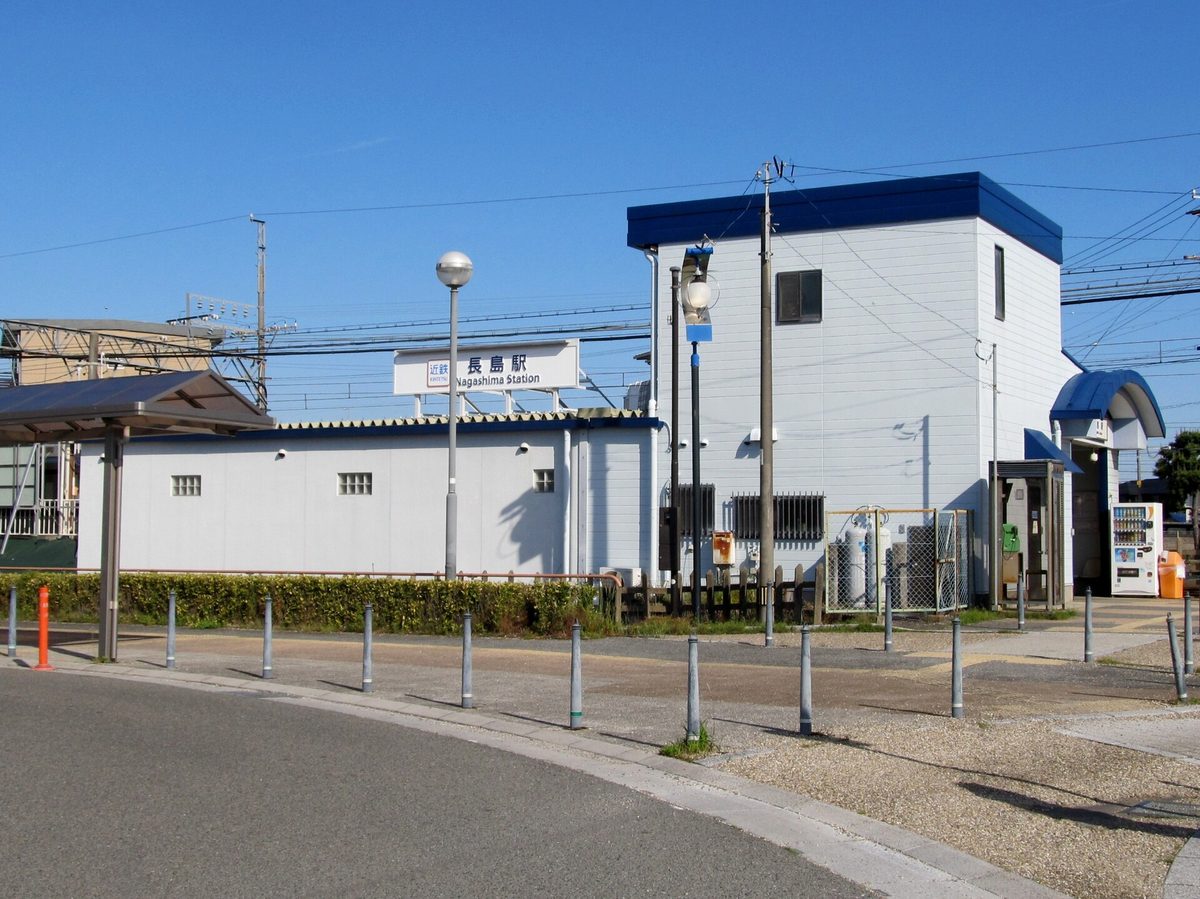 Kintetsu-Nagashima Station building: a small two-storey white station house with blue eaves and a sign reading Kintetsu Nagashima Station, a forecourt with a row of bollards in front and a shelter on the left