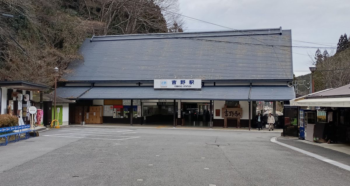 近鐵吉野站站舍正面:灰色大屋頂的木造平房,屋簷下掛著「近鉄 吉野駅 YOSHINO STATION」站名牌,站前是一片柏油廣場,兩側有土產店