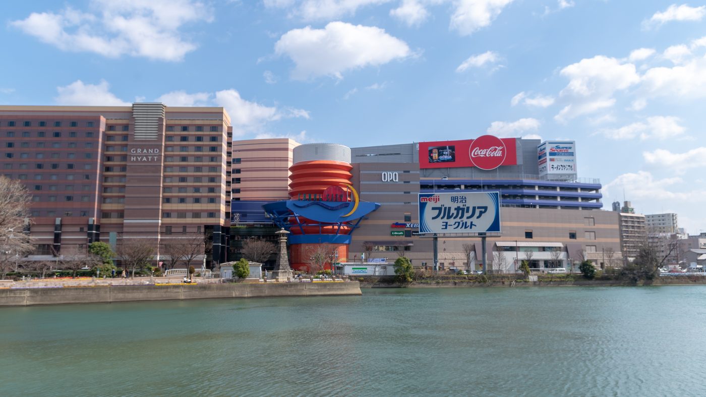 從那珂川對岸看博多運河城：左邊是寫著 GRAND HYATT 的飯店大樓，中間是橘色與藍色的圓弧入口造型，右邊的商場外牆掛著大型廣告看板，前景是河面