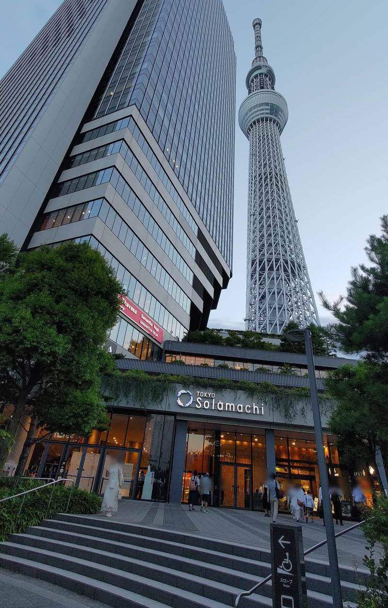 傍晚從階梯下往上看晴空街道 East Yard 的一樓入口：入口上方寫著 TOKYO Solamachi，入口上面是晴空塔東塔的玻璃高樓，右邊是晴空塔，右下角立著指向左邊坡道的牌子