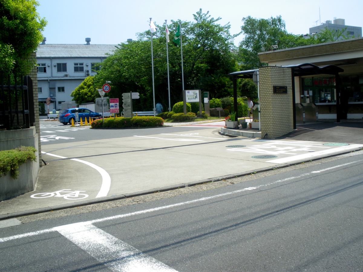 麒麟啤酒橫濱工場的車輛出入口：畫面右側是一座淺黃色磚砌的門柱，柱面嵌著一塊深色金屬銘板，上頭印著 KIRIN 標誌與「キリンビール株式會社 橫濱工場 正門」；門柱右邊是一間開著窗的警衛室，玻璃上貼著紅色大字「總合案內所」，窗前排著幾支不鏽鋼的擋車柱與一段黑色雨遮走廊。左側是一條進廠道路，路口立著限速 20 的圓形標誌、幾個指向廠內各設施的箭頭指標牌與三支旗桿，遠處停著一輛藍色小車，路面上畫著白色的通行標線與一個自行車圖示。晴天，樹很綠