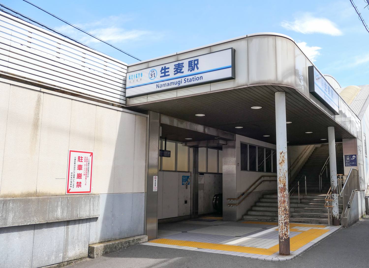 京急線生麥站的東口出入口：一棟灰白色的站體，屋簷下掛著藍白配色的橫式站名燈箱，左端是 KEIKYU 京急電鐵標誌與藍圈裡的車站編號 KK31，中央大字寫著「生麦駅」，下面一行英文 Namamugi Station；出入口是一個沒有閘門的開放式穿堂，裡面看得到一道通往月台的水泥樓梯與扶手、天花板嵌燈，以及地面上的黃色導盲磚，右側牆上另有一塊寫著電梯的指示牌。左邊白色外牆上貼著一張紅框直式告示，大字寫著「駐車厳禁」。晴天，畫面裡沒有人