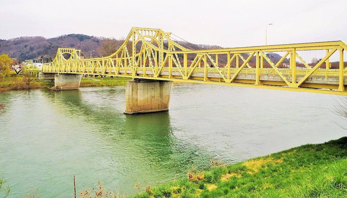 橫跨北上川的珊瑚橋：整座橋是黃色的鋼桁架，兩座橋墩上各有一段拱起的塔狀桁架，河水偏綠，橋的右端後方看得到一小串鯉魚旗，對岸的樹還是紅褐色
