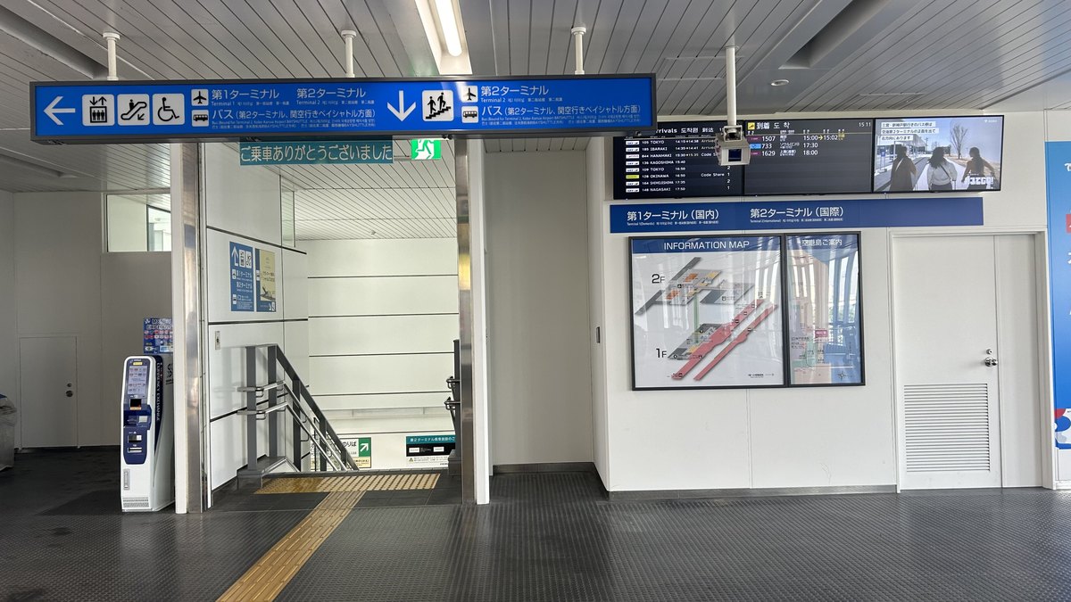 Port Liner 神戸空港站 1 樓出口:天花板掛著藍色指標,左箭頭寫第1ターミナル、第2ターミナル,下箭頭寫第2ターミナル與「バス(第2ターミナル、関空行きベイシャトル方面)」,下方是往下的樓梯;右側牆上有航班到站看板與樓層導覽圖