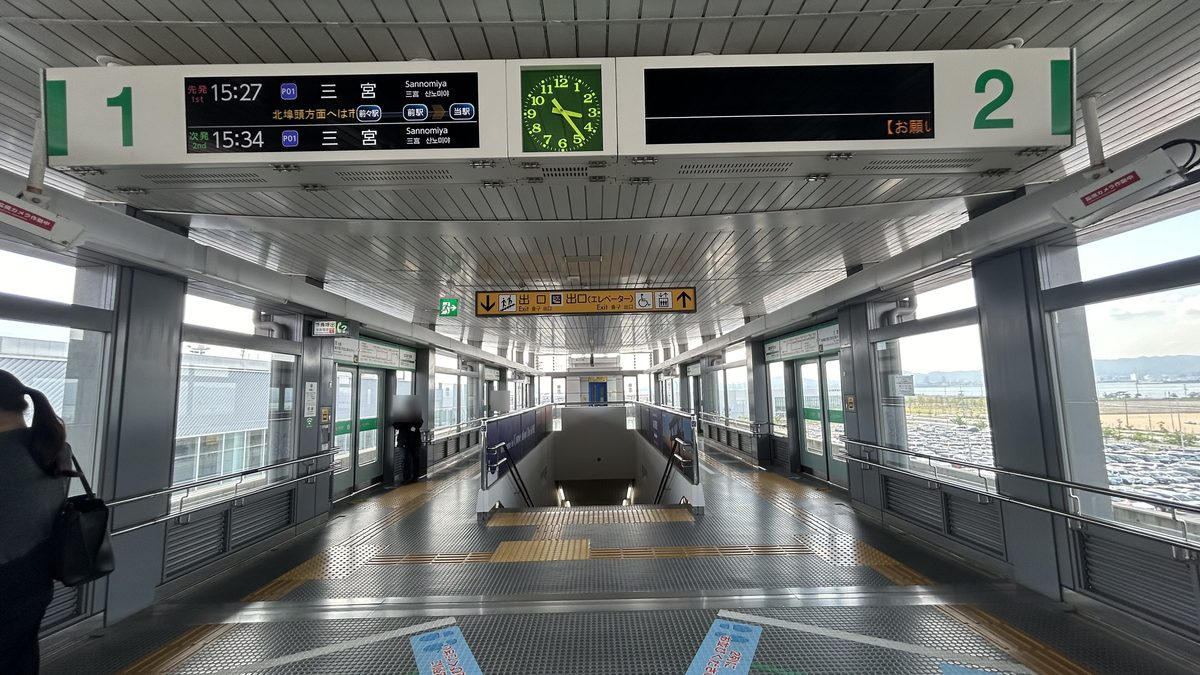 Port Liner 神戸空港站月台:天花板掛著 1 號、2 號線發車看板,1 號線顯示先發 15:27、次發 15:34 往三宮,中間是綠色時鐘;兩側是月台門,中間樓梯往下通往出口,右邊窗外看得到停車場與海