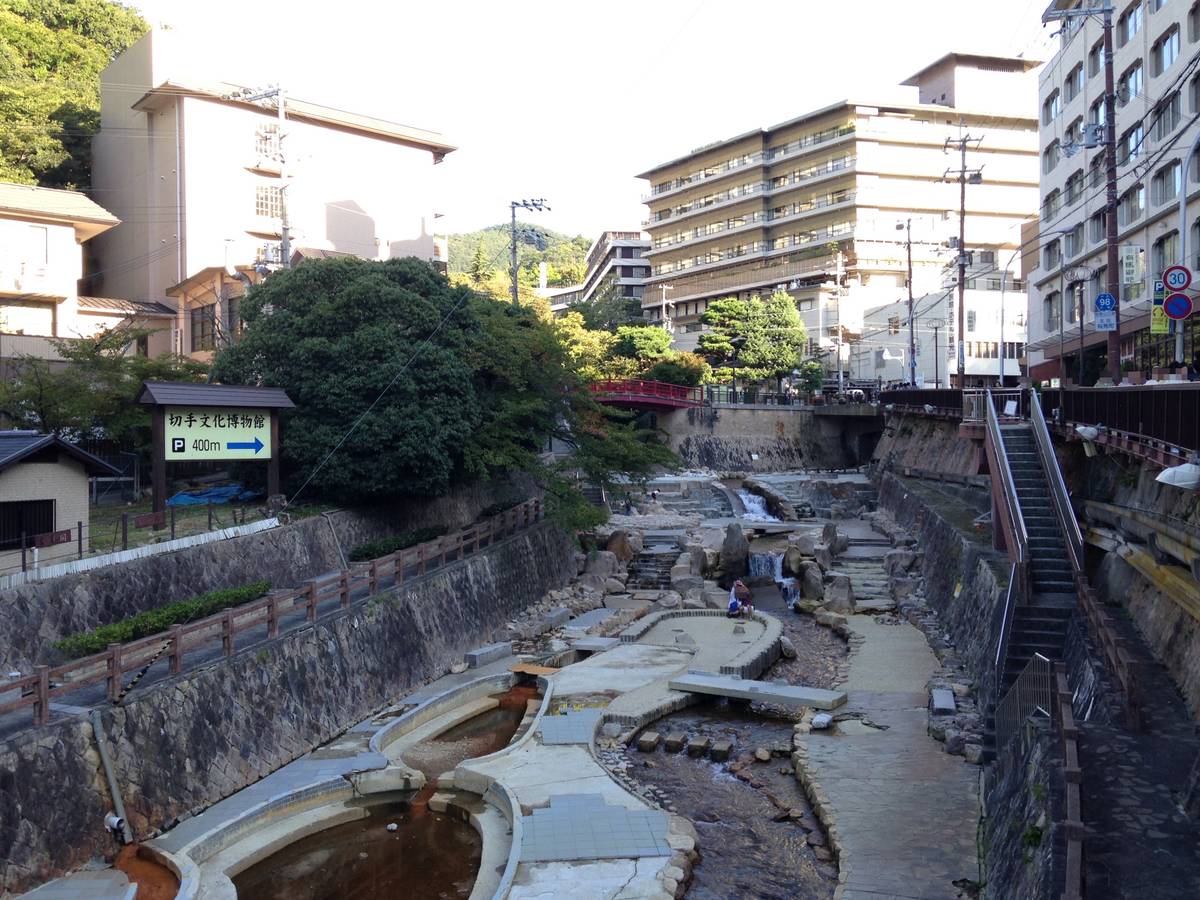 從有馬溫泉太閤橋往上游看的有馬川:石砌護岸之間是整理成親水步道的河床,河水帶著溫泉的紅褐色,遠處有一座紅色欄杆的小橋,兩岸是溫泉旅館大樓與山坡上的綠樹,左邊路旁有「切手文化博物館 400m」的指示牌