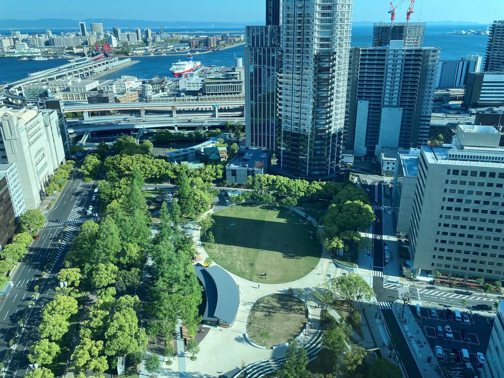 Higashi Yuenchi Park in Kobe seen from above: trees around one large and one small round lawn, a building with a curved grey roof beside them, and towers, an elevated road and the port beyond