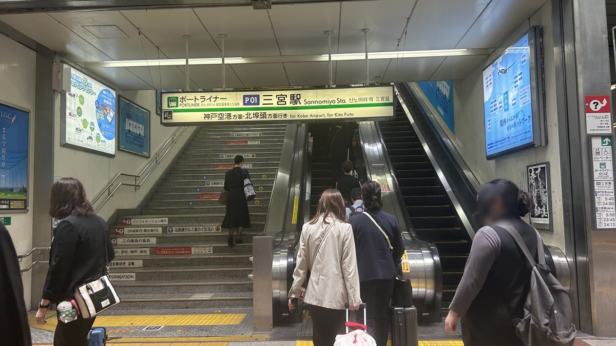 Port Liner 三宮站入口:上方招牌寫「ポートライナー P01 三宮駅」與「神戸空港方面・北埠頭方面行き」,下面是並排的樓梯與兩座手扶梯,幾位旅客拉著行李箱往上走;右邊柱子上的看板列出往神戸空港方向首班 5:40、末班 24:00
