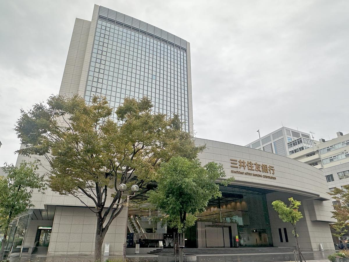 The Sumitomo Mitsui Banking Corporation Kobe headquarters on an overcast day: a curved entrance canopy with gold lettering, a glass tower behind it, and steps and street trees in front