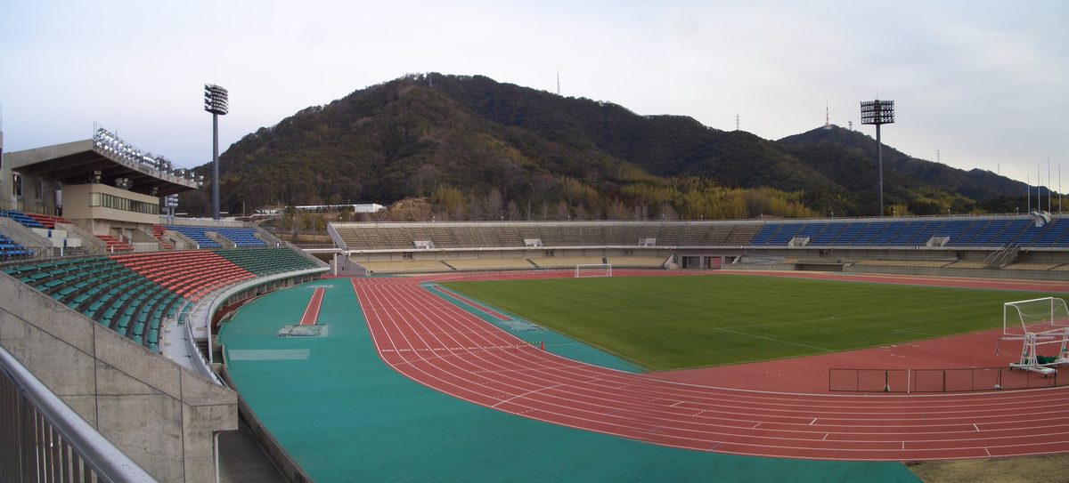 從看台高處看春野綜合運動公園陸上競技場:紅色的田徑跑道圍著綠色草皮,左邊是有屋頂的主看台與照明塔,對面是一長排藍色座椅的看台,背後緊貼著一座長滿樹的山