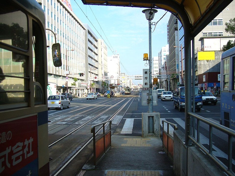 從路面電車県庁前電停的月台往東看:月台中央的站牌寫著「県庁前」,左邊停著一輛電車,路中間兩條電車軌道往前延伸,兩旁是辦公大樓與車道上的汽車