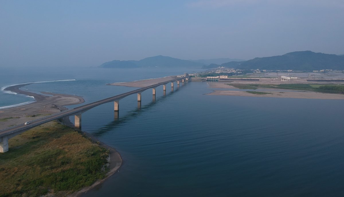 從高處俯瞰仁淀川河口:一座細長的多跨水泥橋從左下角斜斜橫過寬闊的河口,左邊是沙洲與太平洋的浪花,對岸是平地上的房子與一排山