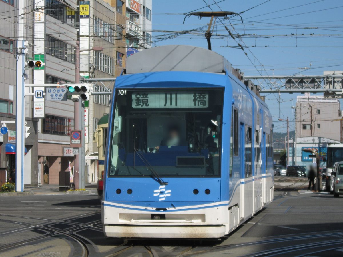 一輛藍白色的低底盤路面電車迎面開來,車頭上方的行先表示寫著「鏡川橋」,左邊交通號誌旁的路牌寫著「はりまや交差点」,頭上有電車架線