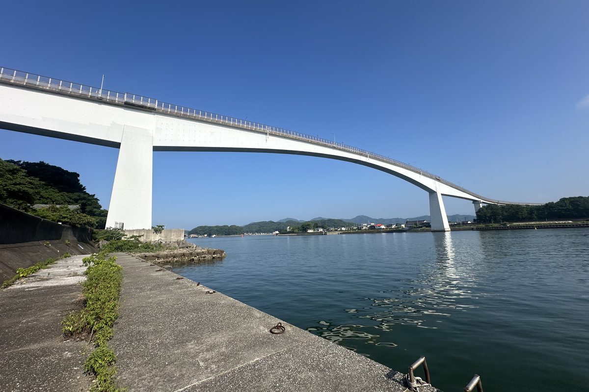 從浦戶灣岸邊的水泥堤防仰望浦戶大橋:白色橋身從左上方高處的橋墩一路往右下降到對岸,中段離水面很高,橋下是平靜的海灣,對岸是低矮的山與房子