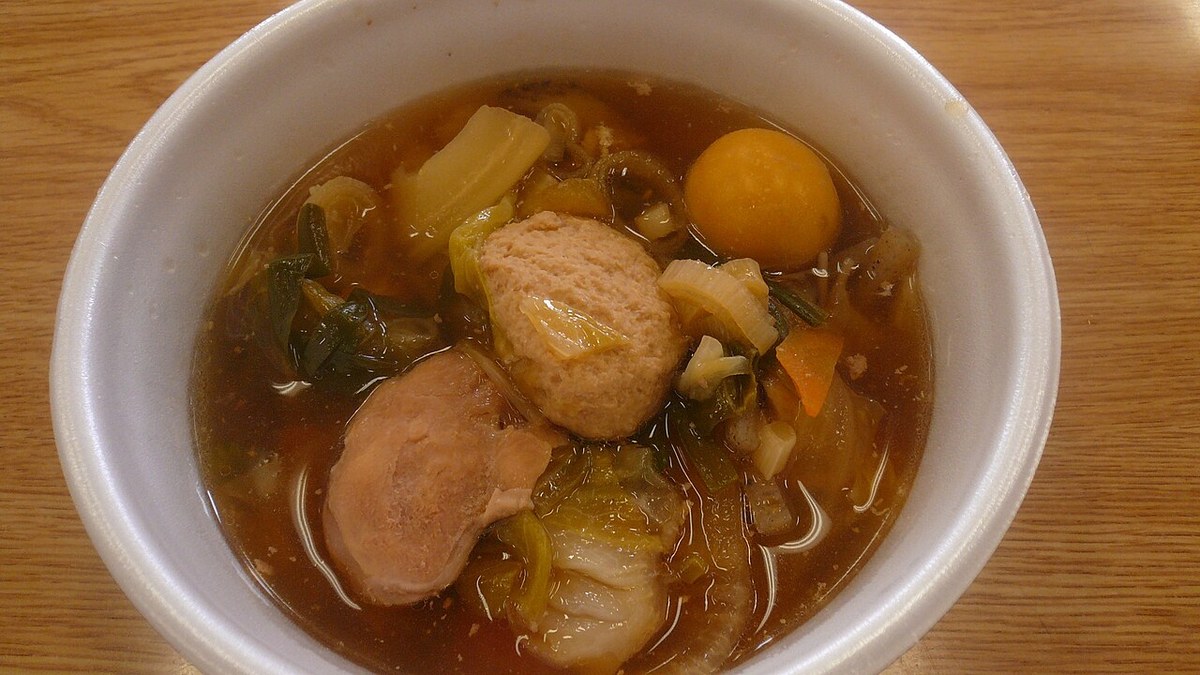 Close-up of a bowl of chanko nabe: a piece of chicken, a chicken meatball, napa cabbage, carrot, spring onion and one round orange item in a brown broth, in a white disposable bowl on a wooden table