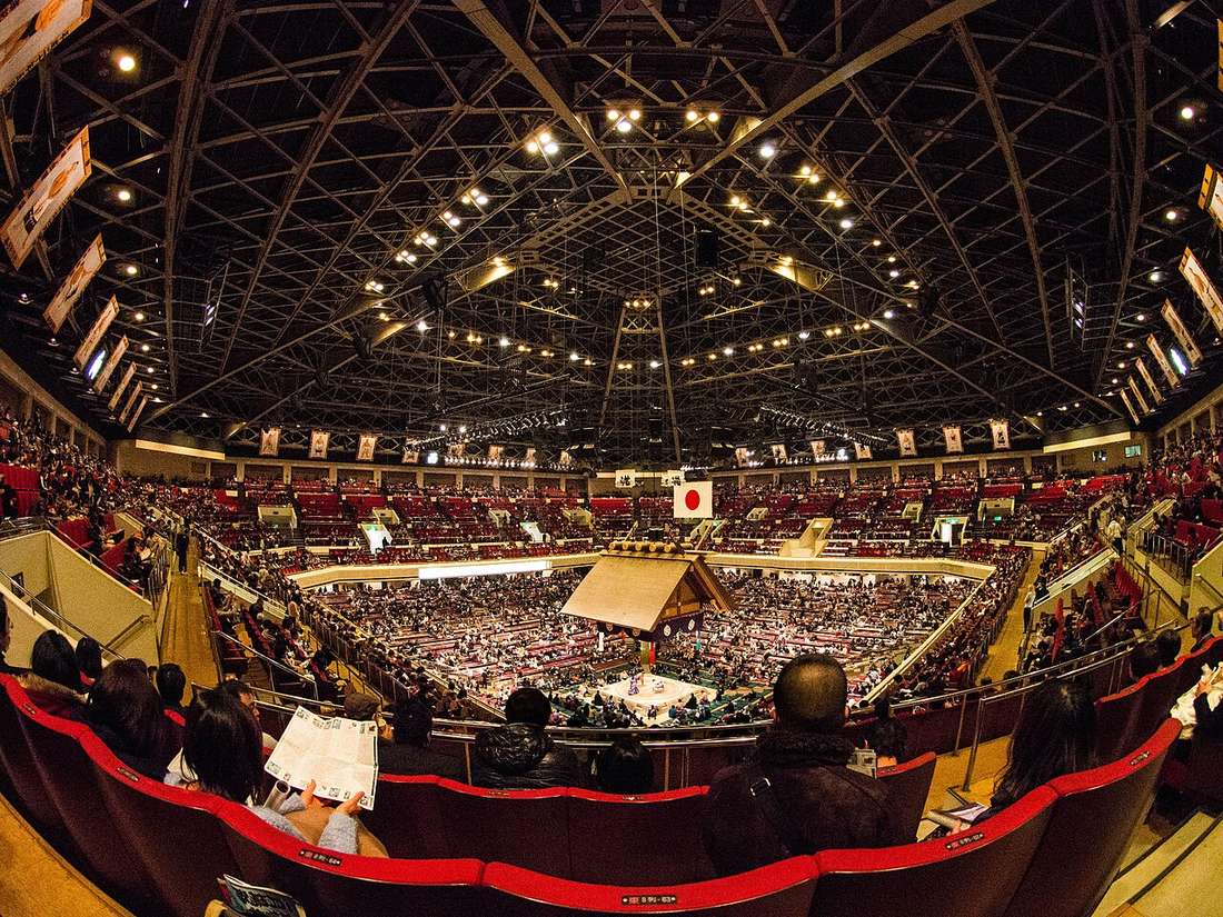 Wide fisheye view down from a second-floor chair seat inside the Kokugikan: the ring under its suspended wooden roof at the centre, a grid of ground-floor box seats around it, a ring of red chair seats above, steel roof trusses and floodlights overhead, and a Japanese flag with framed champion portraits on the far wall