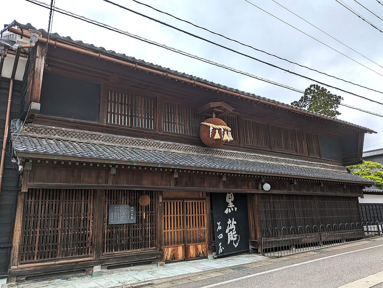 The Kokuryu Shuzo brewery shop in Eiheiji, Fukui prefecture: a two-storey dark timber townhouse, a straw cedar ball hung with a shimenawa rope beneath the small roof over the lattice windows, a black sign in Japanese calligraphy beside the entrance, and a black notice board with vertical Japanese text on the wall to the left.