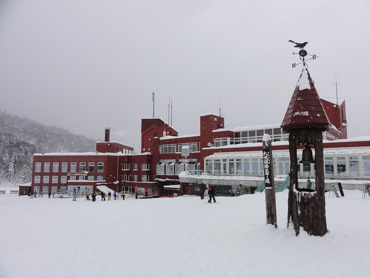 The base of Sapporo Kokusai during snowfall, the dark red centre building under deep roof snow, skiers scattered on the nursery slope in front and a wooden bell tower on the right carrying a vertical Sapporo Kokusai Ski Resort sign