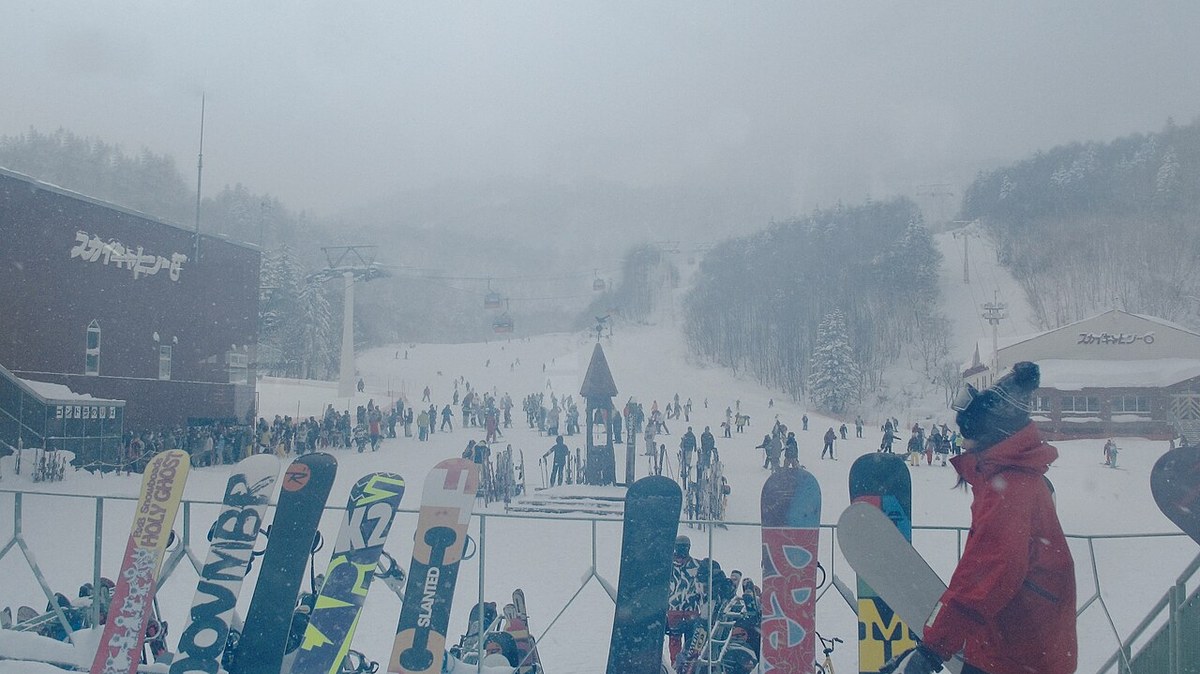 The base of Sapporo Kokusai in falling snow, boards and poles stacked along a railing in the foreground, crowds of skiers on the lower slope, Sky Cabin signage on the buildings to left and right and gondola cabins climbing into the mist
