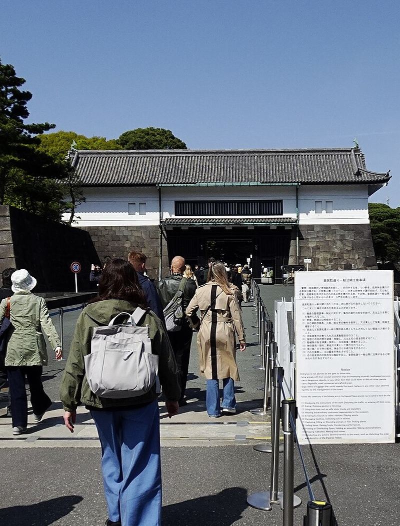 乾通一般公開期間的皇居坂下門：遊客背對鏡頭沿著圍欄柱往門洞走，門是石垣上一座白牆黑瓦的櫓門，右側立著一塊寫滿注意事項的白色告示板