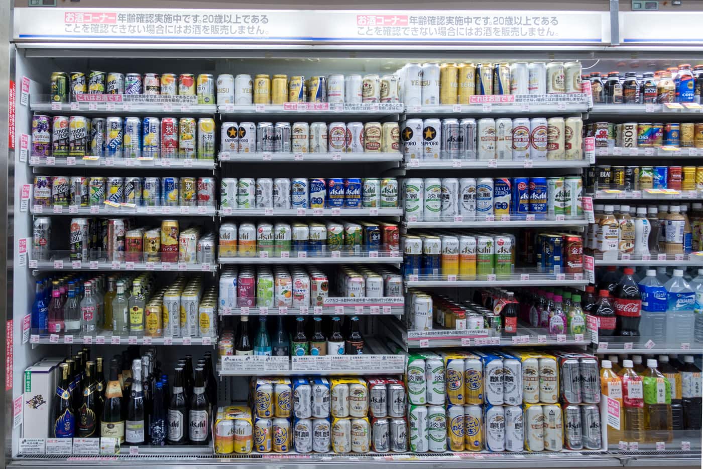 Full view of the alcohol cooler in a Japanese convenience store: a red banner along the top reads the Japanese age-verification notice, the left-hand columns hold canned chuhai, and the centre and right shelves hold canned beer, happoshu and new-genre cans, each row edged with small pink price labels.