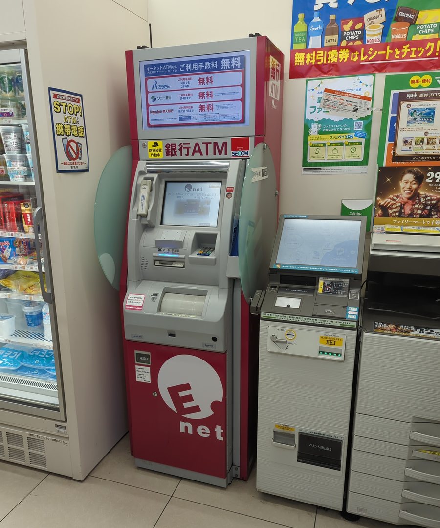 A red E-net bank ATM standing against the wall inside a FamilyMart, its cabinet carrying the E-net logo and a return slot, an illuminated panel above listing fee-free conditions for three Japanese banks, and an anti-fraud poster on the wall warning against mobile phone use at the ATM; to its right stands a multi-copy machine with a touchscreen, a coin slot and a print output tray