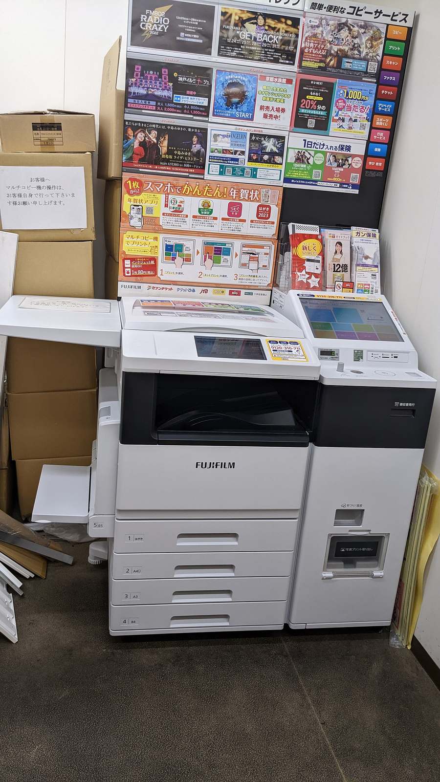 A white FUJIFILM multi-copy machine in the corner of a 7-ELEVEN, joined on the right by a payment unit with its own touchscreen and coin slot; the wall behind is covered in concert and exhibition flyers, with a black service board listing copy, print, scan, fax, tickets and insurance; stacked cardboard boxes on the left carry a handwritten notice