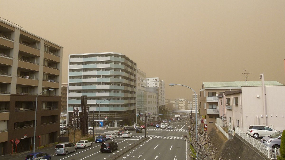 A Yokohama crossroads and dual carriageway flanked by apartment blocks; the whole sky is a flat yellow-brown with no cloud shapes, and the more distant towers have dissolved into pale grey silhouettes