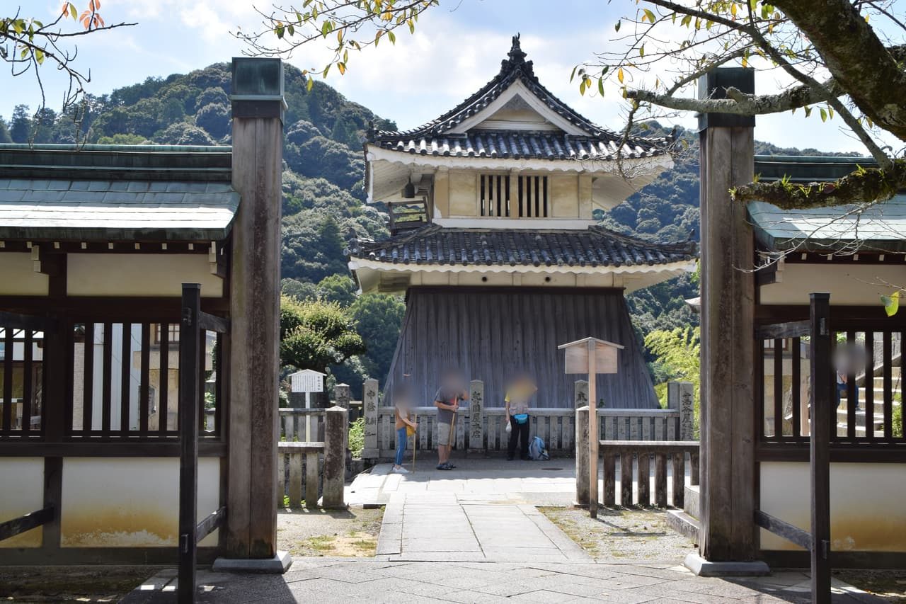 Kotohira-gu's drum tower seen between wooden gateposts: two tiled roofs over a flared plank base, with three visitors by the stone fence in front